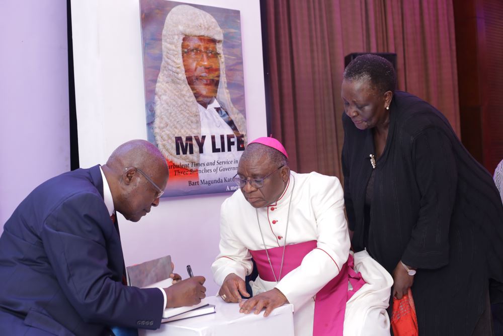 Former Chief Justice Bart Katureebe autographs The Arch Bishop of Kampala Paul Semogerere as Former Vice President Specioza Kazibwe looks on (PHOTO/Courtesy)